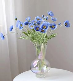 blue flowers are in a clear vase on a white table next to a curtained window