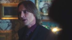 a man with long hair wearing a suit and tie talking to another man in a room