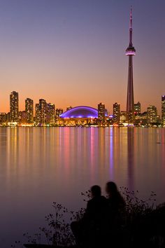 two people sitting on a bench looking out over the water in front of a city skyline