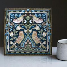 a decorative tile with birds and flowers on it next to a white cup that is sitting on a table