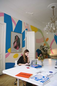 a woman sitting at a table with a laptop in front of her and paintings on the wall behind her