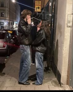 two people standing on the sidewalk talking on their cell phones while one person holds his hand up to his mouth