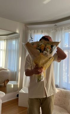 a man holding flowers in front of his face while standing in a living room next to a window