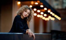 a woman with curly hair is sitting on a ledge in front of some string lights