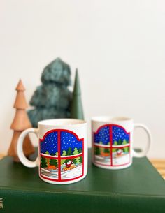 two coffee mugs sitting on top of a green book next to a christmas tree