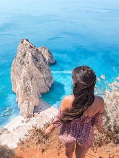 a woman standing on top of a cliff next to the ocean with her hair blowing in the wind