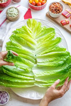 two hands are holding lettuce leaves on a white plate with other food and condiments