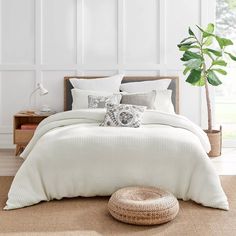 a bed with white sheets and pillows next to a potted plant on the floor