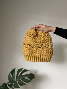 a hand holding a yellow crocheted hat on top of a table next to a potted plant