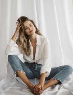 a woman sitting on top of a bed wearing ripped jeans and a white button up shirt