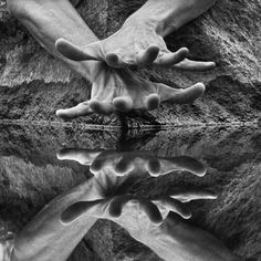 two hands reaching out towards each other in front of water and rocks with their reflection on the ground