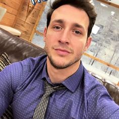 a man in a blue shirt and tie sitting on a couch with his arms crossed