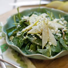 a salad with cheese and greens in a green bowl on a wooden table next to utensils