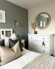 a white bed topped with pillows next to a dresser under a round mirror in a bedroom