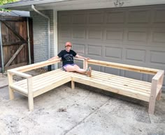 a man sitting on top of a wooden bench in front of a garage with his legs crossed