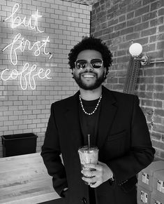 a man wearing sunglasses holding a drink in front of a brick wall with the words but first coffee written on it