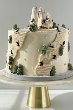 a cake with frosting decorated with trees and skiers on the top is sitting on a gold pedestal