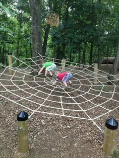 two children playing in a large spider web at the park with trees and bushes behind them