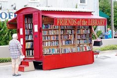 a man standing in front of a red book stand with books on it and people walking by