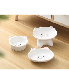 three white bowls with cats on them sitting on a wooden floor next to a plant