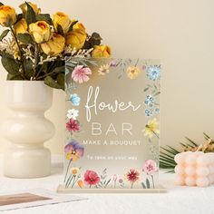 a flower bar sign sitting next to a vase filled with yellow and pink flowers on top of a white table