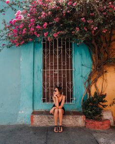 a woman sitting on a ledge in front of a blue building with pink flowers growing over it