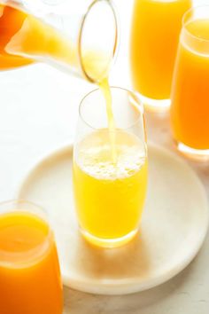 orange juice being poured into a glass on a white plate with other glasses in the background