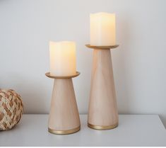 two white candles sitting on top of a table next to a basket and lamp shade