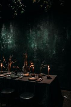 a long table with candles and plates on it in front of a dark wall, surrounded by greenery