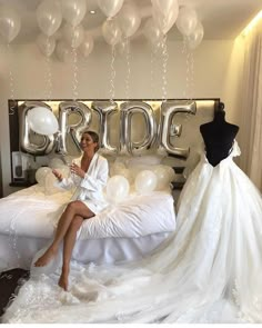 a woman sitting on top of a bed next to a wedding dress and balloon arch