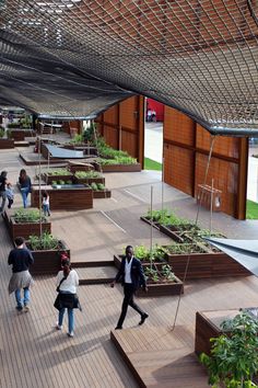 people are walking around an outdoor garden area with raised planters and wooden benches on the floor