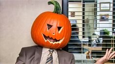 a man in a suit and tie with a pumpkin on his head, sitting at a table