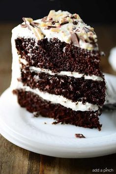 a slice of chocolate cake with white frosting and chopped nuts on top, sitting on a plate