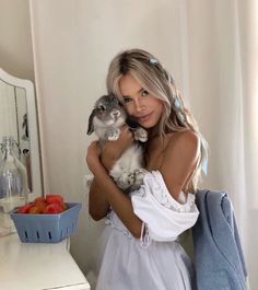 a woman holding a small kitten in her arms while standing next to a table with fruit on it