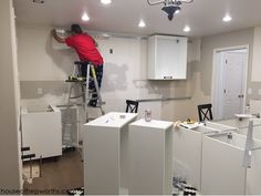 a man on a ladder painting the walls in a room with white cabinets and appliances