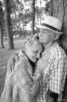 an old man and woman embracing each other in front of a tree with pine trees behind them