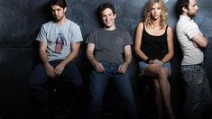 four people sitting on stools in front of a black wall and one person leaning against the wall