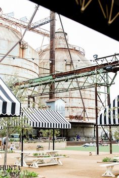 an industrial area with tables and umbrellas in the foreground, near large tanks