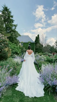 a woman in a white wedding dress walking through a garden with purple flowers and greenery