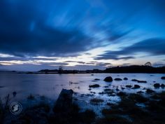 the sky is dark and cloudy over the water with rocks in the foreground at dusk