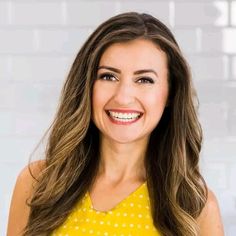 a woman with long brown hair and yellow top smiling at the camera in front of a white brick wall