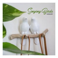 two white birds sitting on top of a wooden branch next to green leaves and the words singing birds hanger