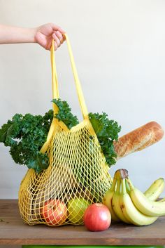 a person holding a yellow mesh bag with fruits and vegetables in it next to some bread