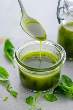 a spoon full of green liquid being poured into a glass jar with basil leaves on the side