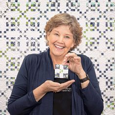 an older woman holding up a small piece of paper in front of a quilted wall