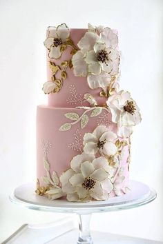 a pink and gold wedding cake with white flowers on the top tier is sitting on a glass stand