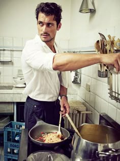 a man standing in a kitchen preparing food on top of a frying pan with a wooden spoon