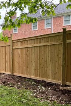 a wooden fence in front of a red brick house with grass and dirt on the ground