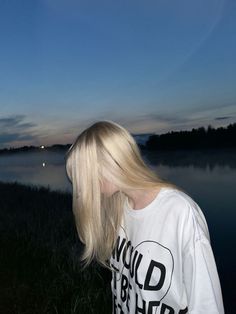 a blonde woman with long hair standing in front of the water at dusk looking off into the distance