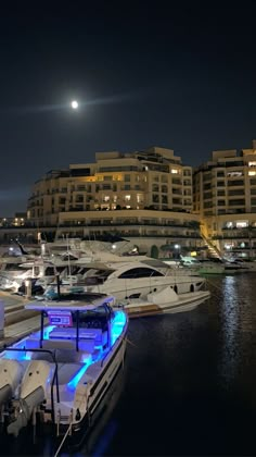 several boats are docked in the water at night time, with city lights and buildings in the background